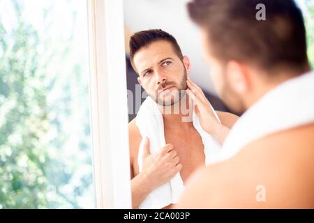 Nahaufnahme eines hübschen jungen Mannes mit einem Handtuch im Nacken, der sein Gesicht im Badezimmerspiegel bewundert. Stockfoto