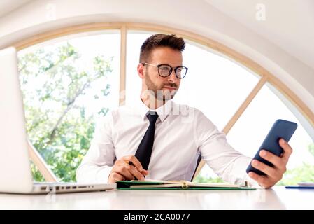 Geschäftsmann, der am Schreibtisch hinter seinem Laptop sitzt und SMS-Nachrichten mitgibt. Heimbüro. Stockfoto