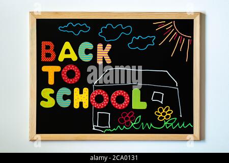 Tafel mit Text Zurück zur Schule mit bunten Holzbriefen und Kreidezeichnung von Schulhaus, Gras, Blumen, Sonne und Wolken. Worte Zurück zur Schule Stockfoto