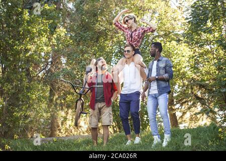 Junge BMX-Fahrer in Gesellschaft mit seinen multiethnischen Freunden zu Fuß, um eine gute Zeit im Stadtpark haben, glückliche junge Frau huckbacking auf ihrem Freund Stockfoto