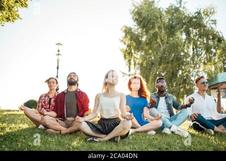 Yoga im Park. Interracial Freunde meditieren und verbringen ihre Freizeit im Freien. Konzept von gesunder Lebensweise, Freundschaft und Vielfalt. Panarom Stockfoto