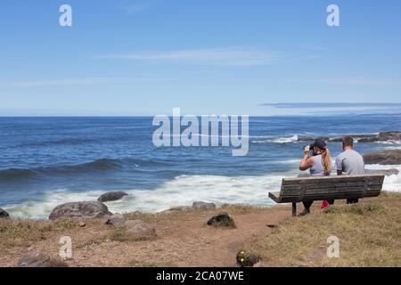 Ein Mann und eine Frau mit Gesichtsmasken sitzen auf einer Bank am Meer in Yachats, Oregon, USA. Stockfoto