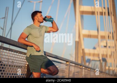 Der junge Mann trainiert draußen auf der Brücke in der Stadt. Er trinkt Wasser. Stockfoto