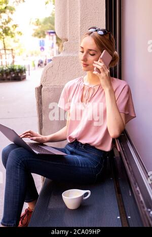 Porträtaufnahme einer jungen Frau, die einen Anruf hat und einen Laptop benutzt, während sie im Café sitzt. Stockfoto