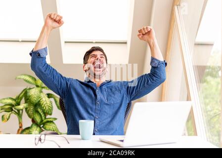 Fröhlicher Geschäftsmann feiert mit erhobenem Arm in der Luft, während er am Schreibtisch in seinem Heimbüro sitzt und an einem Laptop arbeitet. Heimbüro. Stockfoto