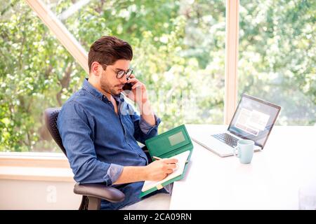 Aufnahme eines Geschäftsmannes, der einen Anruf macht und etwas schreibt, während er hinter seinem Laptop am Schreibtisch sitzt. Stockfoto
