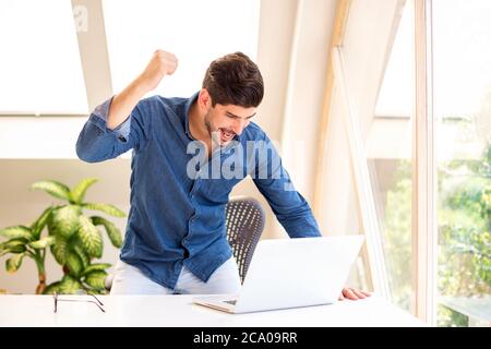 Fröhlicher Geschäftsmann feiert mit erhobenem Arm in der Luft, während er an einem Schreibtisch in seinem Heimbüro steht und an einem Laptop arbeitet. Heimbüro. Stockfoto