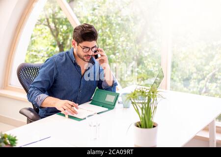 Aufnahme eines Geschäftsmannes, der einen Anruf macht und in seinen Ordner schreibt, während er am Schreibtisch sitzt. Stockfoto