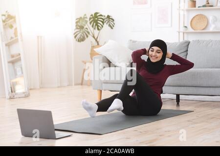 Sportliche arabische Frau in Hijab tun abs-Übungen mit Laptop zu Hause Stockfoto