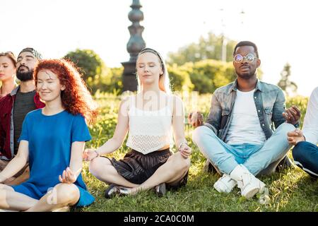 Vielfältige Yoga-Klasse von fröhlichen jungen Freunden im Sommer Casual Wear sitzen in Lotus-Pose auf grünen lown im öffentlichen Park, Vorderansicht mit Fokus auf fe Stockfoto