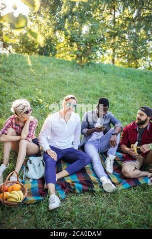 Multirassische Gruppe von sorglosen Menschen in lässigen Sommer tragen sitzen auf Park lown und beobachten etwas intresting in Smartphone gekleidet. Frauen und Männer Stockfoto