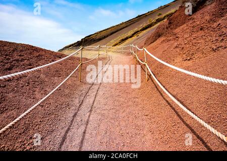 Einsamer Fußweg durch eine atemberaubende rote vulkanische Felslandschaft unter blauem Himmel, gesehen in El Golfo, Lanzarote, Kanarische Inseln, Spanien. Stockfoto