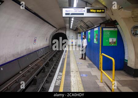 Ein Mann, der eine Gesichtsbedeckung trägt, wartet auf einem leeren Bahnsteig auf einen Zug auf einer Londoner U-Bahn-Station nach der Sperre. London, Großbritannien Stockfoto