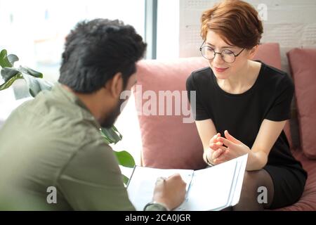 Junge Frau sitzt auf der Couch und spricht mit männlichen indischen Psychologen, während Arzt Notizen macht Stockfoto