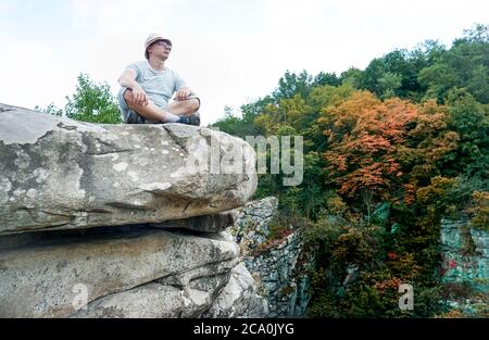 Ein Kerl sitzt auf einem Felsen in den Bergen auf einem Stein, gekleidet in ein T-Shirt und Shorts Stockfoto