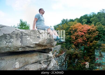 Ein Kerl sitzt auf einem Felsen in den Bergen auf einem Stein, gekleidet in ein T-Shirt und Shorts Stockfoto