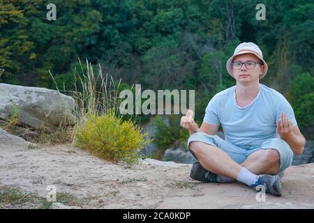 Ein Kerl sitzt auf einem Felsen in den Bergen auf einem Stein, gekleidet in ein T-Shirt und Shorts Stockfoto