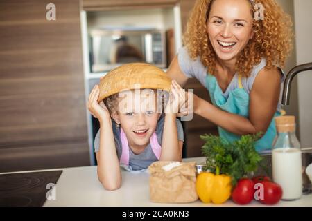 Schöne Mutter und nette kleine Tochter Spaß in der Küche, während die Vorbereitung Muffins in der Schüssel kochen. Glückliches Mädchen trägt die Schüssel auf dem Kopf und Stockfoto