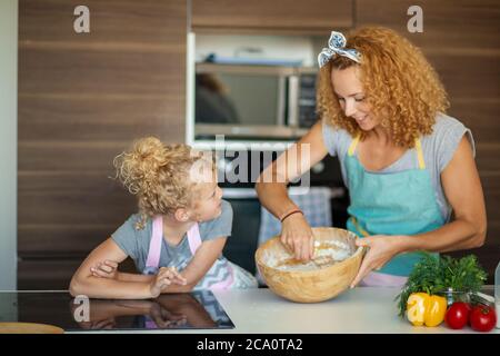 Kaukasische Mutter und kleine emotionale Mädchen knacken Eier zu Mehl für Teig zum Geburtstag des Vaters.. Mutter Lehrer ihre Tochter, wie man selbst gemacht Stockfoto