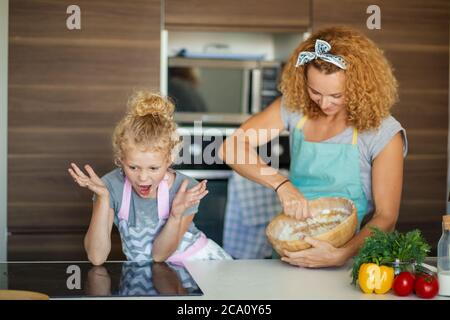 Kaukasische Mutter und kleine emotionale Mädchen knacken Eier zu Mehl für Teig zum Geburtstag des Vaters.. Mutter Lehrer ihre Tochter, wie man selbst gemacht Stockfoto