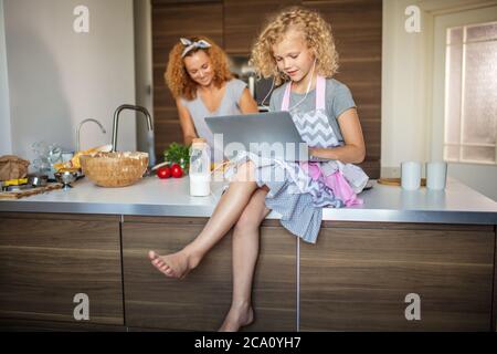 Nettes lächelndes Mädchen mit Laptop und Kopfhörer sitzen auf dem Küchentisch, Rezept zu ihrer Mutter, die Familie Abendessen macht lesen. Stockfoto