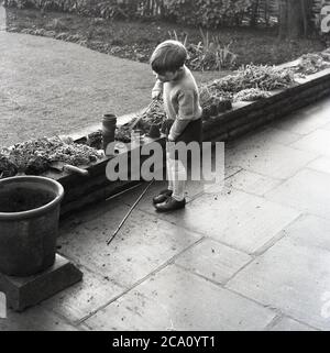 1960s, historisch, Beginn Gartenarbeit jung,...ein kleiner Junge in seiner Grundschule Uniform draußen auf einer Terrasse spielen mit einer Reihe von kleinen Steingut oder Terrakotta-Töpfe, nachdem gerade den Boden auf die Seite des Blumenbeet entleert, England, Großbritannien. Stockfoto