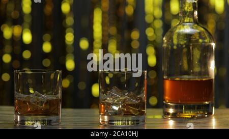 Goldener Whiskey, Cognac oder Brandy in zwei Gläsern mit Eiswürfeln und Flasche auf Holztisch. Glänzend Gold Party Feier Hintergrund. Gläser Alkohol trinken Bourbon, Rum Stockfoto