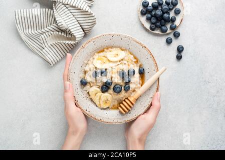 Weibliche Hände halten Schüssel Haferbrei Haferbrei mit Bananenscheiben, Heidelbeeren, Kokosflocken und natürlichen rohen Honig über grauen Beton Hintergrund, Tab Stockfoto