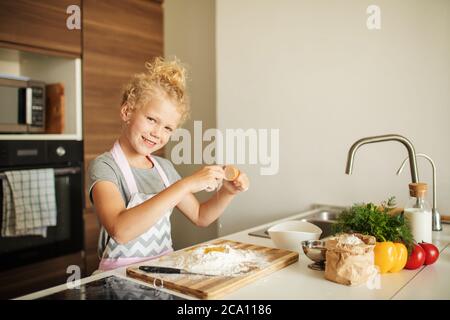 Kleine Köchin steht in der Küche, schaut auf die Kamera und knackt Eier bis Mehl, was für Eltern Überraschung macht, Pizza kocht. Stockfoto