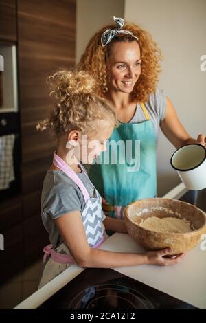 Schöne kaukasische Mutter und Tochter Spaß in der Küche, Mutter Gießen Milch, während Tochter hält Schüssel mit Mehl. Küchenangelegenheiten. Stockfoto