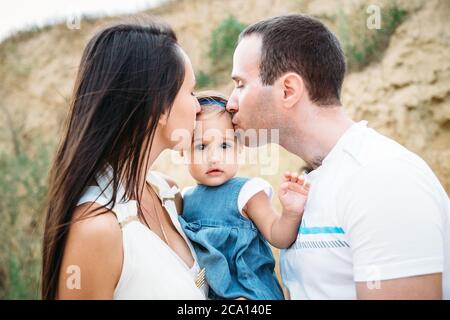 Nahaufnahme der jungen Familie in weißen T-Shirts mit einer kleinen Tochter in Kleid Umarmung und küssen, im Freien Hintergrund Stockfoto