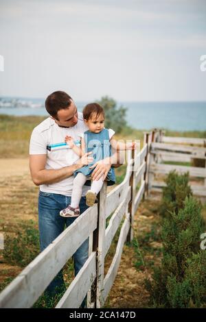 Nahaufnahme von kleinen Mädchen in blauen Jeans Kleid sitzt auf einem Zaun in der Umarmung des Vaters Stockfoto