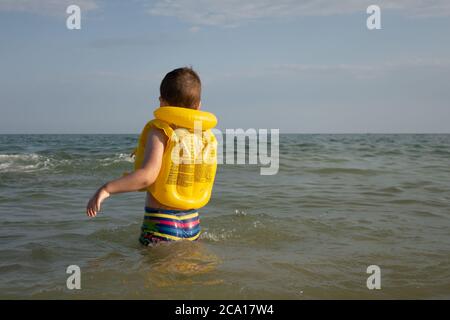 Ein 5-jähriger Junge mit gelber Schwimmweste und raingenbogenfarbenen Shorts betritt das Meer. Stockfoto