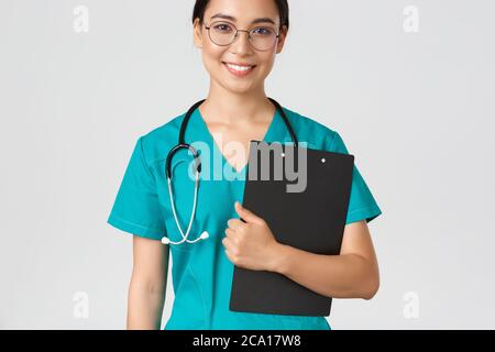 Covid-19, Coronavirus-Krankheit, Konzept für Mitarbeiter im Gesundheitswesen. Lächelnd zuversichtlich asiatische Krankenschwester, Arzt in Brille und Scrubs halten Zwischenablage Stockfoto