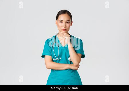 Covid-19, Mitarbeiter im Gesundheitswesen, Pandemiekonzept. Nachdenkliche Ärztin, asiatische Krankenschwester in Scrubs Blick neugierig auf Kamera, denken, suchen Stockfoto