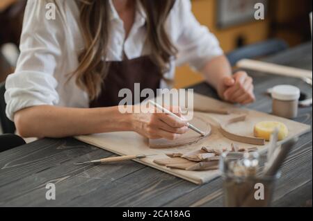 Handwerkerhände verbinden die Tonteile aus rohem Ton, Meisterfinger arbeiten mit Tonstücken, Weibchen sitzt in einer Werkstatt hinter dem Tisch Stockfoto