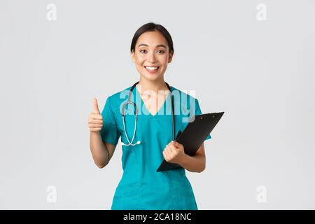 Mitarbeiter im Gesundheitswesen, Virenprävention, Quarantänekampagne Konzept. Lächelnd ziemlich asiatische Arzt, weibliche Praktikantin in Scrubs zeigen Daumen-up, halten Stockfoto