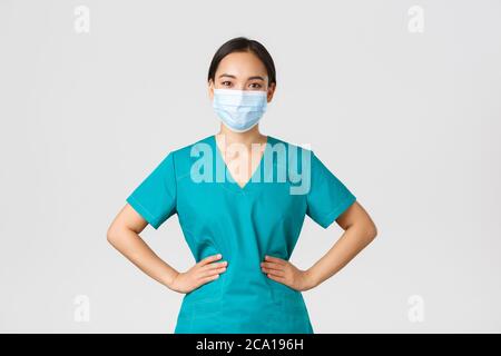 Covid-19, Coronavirus-Krankheit, Konzept für Mitarbeiter im Gesundheitswesen. Fröhlich lächelnd asiatische Ärztin, Arzt in Scrubs und medizinische Maske, Blick optimistisch Stockfoto