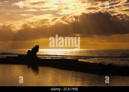 Orangefarbener Sonnenuntergang mit Wolken und Lichtungen von einem Strand mit Felsen im Vordergrund. Romantisches Bild mit Kopierbereich Stockfoto