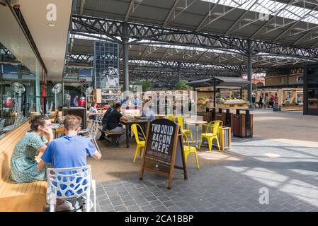 Leute sitzen an Tischen vor einem Restaurant auf dem alten Spitalfields Markt. Der Hauptmarkt und Händler Stände im Hintergrund. London, England, Großbritannien Stockfoto