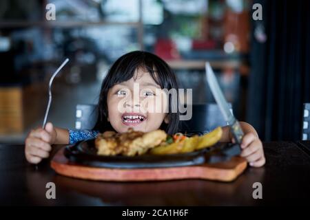 Fröhliches kleines Mädchen, das Steak im Restaurant vor Ort isst Stockfoto