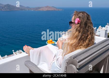 Schöne junge Frau mit Blume im Haar Kaffeetasse sitzen auf der weißen Terrasse Balkon des Hauses oder Hotel mit Meerblick Stockfoto