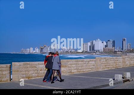 Tel Aviv, Israel - 11. Januar 2017: Drei Freunde spazieren entlang einer Promenade am Mittelmeer, mit der Skyline der Stadt im Hintergrund. Stockfoto