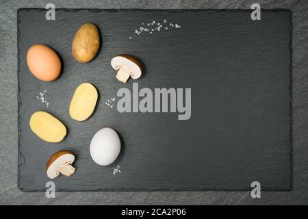 Pilztortilla Zutaten mit zwei halbbraunen Pilzen, einem braunen und einem weißen Ei, halbierten Kartoffeln und groben Salzkörnern auf einer Schieferplatte. Mit Cop Stockfoto