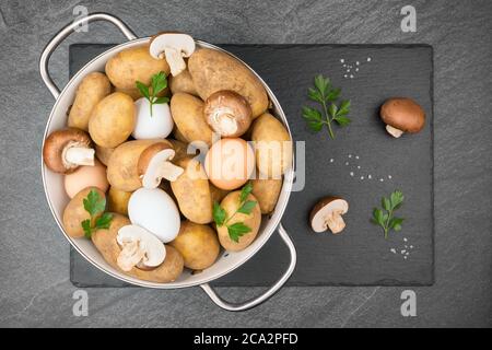 Rohe Kartoffeln, Eier, halbierte und ganze braune Pilze, grobe Salzkörner und Petersilienblätter in einem weißen Emaillesieb auf einem Schieferschlag. Flach liegend. Stockfoto