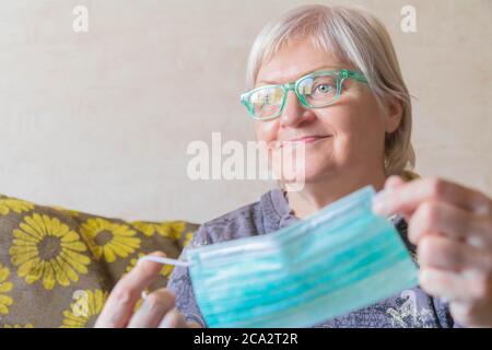 Ältere lächelnde Frau, die zu Hause die Schutzmaske auszieht. Ältere Frau mit Gesichtsmask in den Händen. Lächelnde Dame zieht medizinische Gesichtsmaske nach dem Rückweg aus Stockfoto