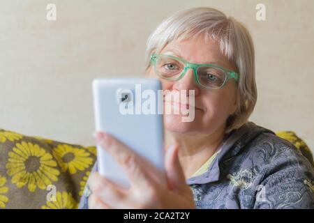 Ältere Frau, die ein Mobiltelefon für die Verbindung mit Verwandten verwendet. Ältere Frau nimmt Selfie. Lady mit Smartphone in der Hand zu Hause. Weiblich, die das Telefon benutzt Stockfoto