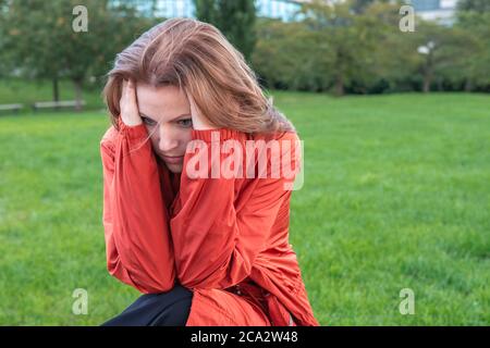 Traurige Frau, die im Park sitzt und den Kopf hält. Depressive Frau allein im Regenmantel im Stadtpark. Frustrierte unglückliche Frau, die an Kopfschmerzen leidet. Verwirrt und Stockfoto