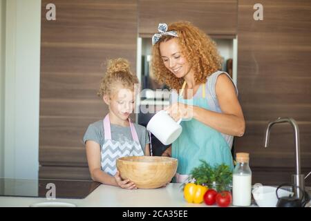 Schöne kaukasische Mutter und Tochter Spaß in der Küche, Mutter Gießen Milch, während Tochter hält Schüssel mit Mehl. Küchenangelegenheiten. Stockfoto