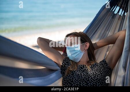Junge Frau trägt medizinische Schutzmaske in einer Hängematte am schönen Meer entspannen. Stockfoto